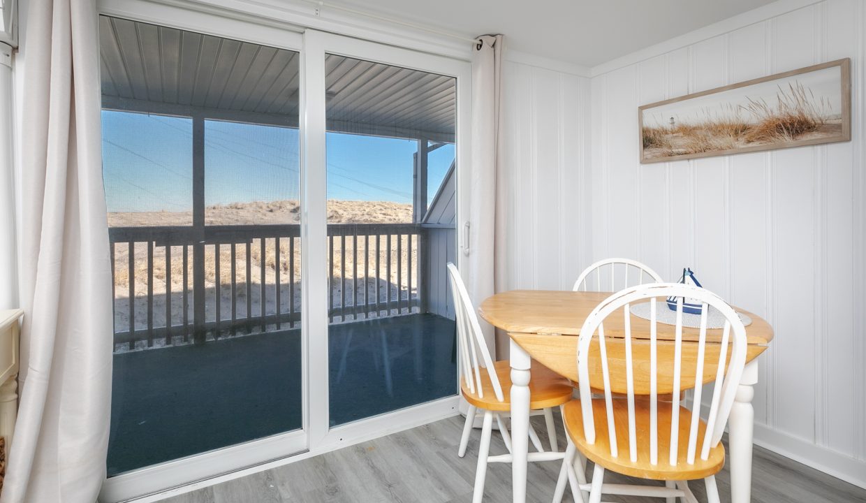 Cozy dining area with a wooden table and white chairs, overlooking a beach landscape through sliding glass doors.