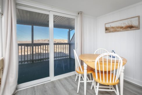 Cozy dining area with a wooden table and white chairs, overlooking a beach landscape through sliding glass doors.