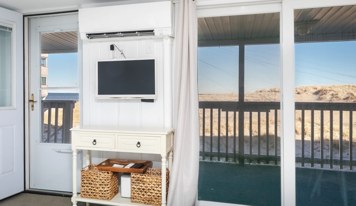 Beach house porch with a flat-screen tv mounted above a white cabinet containing wicker baskets, overlooking sand dunes.