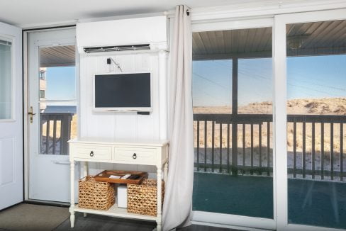 Beach house porch with a flat-screen tv mounted above a white cabinet containing wicker baskets, overlooking sand dunes.