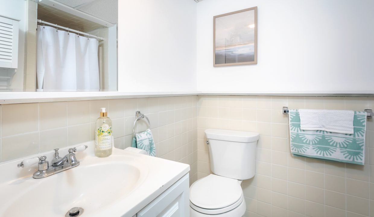 Brightly lit bathroom with a sink, toilet, and shower curtain, featuring a framed picture on the wall and decorative towels.