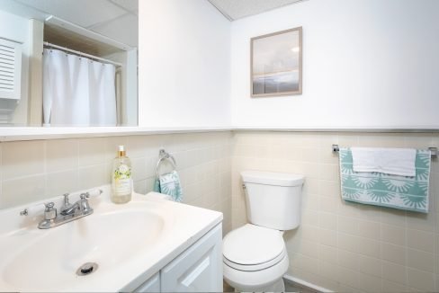 Brightly lit bathroom with a sink, toilet, and shower curtain, featuring a framed picture on the wall and decorative towels.