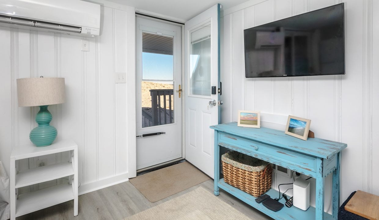 A bright, coastal-styled room with a flat-screen tv mounted above a blue console table, a small desk with a laptop, an open doorway leading to an outdoor area, and a wall-mounted air conditioning unit.