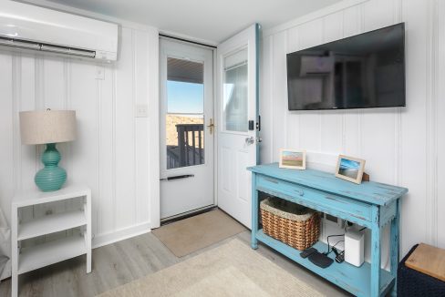 A bright, coastal-styled room with a flat-screen tv mounted above a blue console table, a small desk with a laptop, an open doorway leading to an outdoor area, and a wall-mounted air conditioning unit.