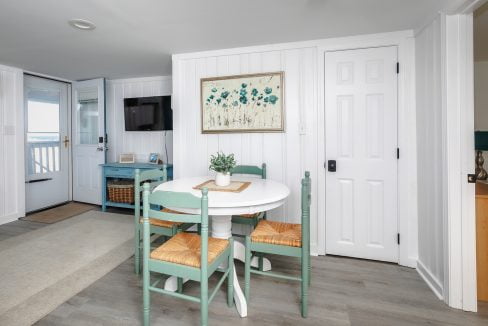 Bright coastal-themed dining area with a white round table, green chairs, and a floral wall art piece.