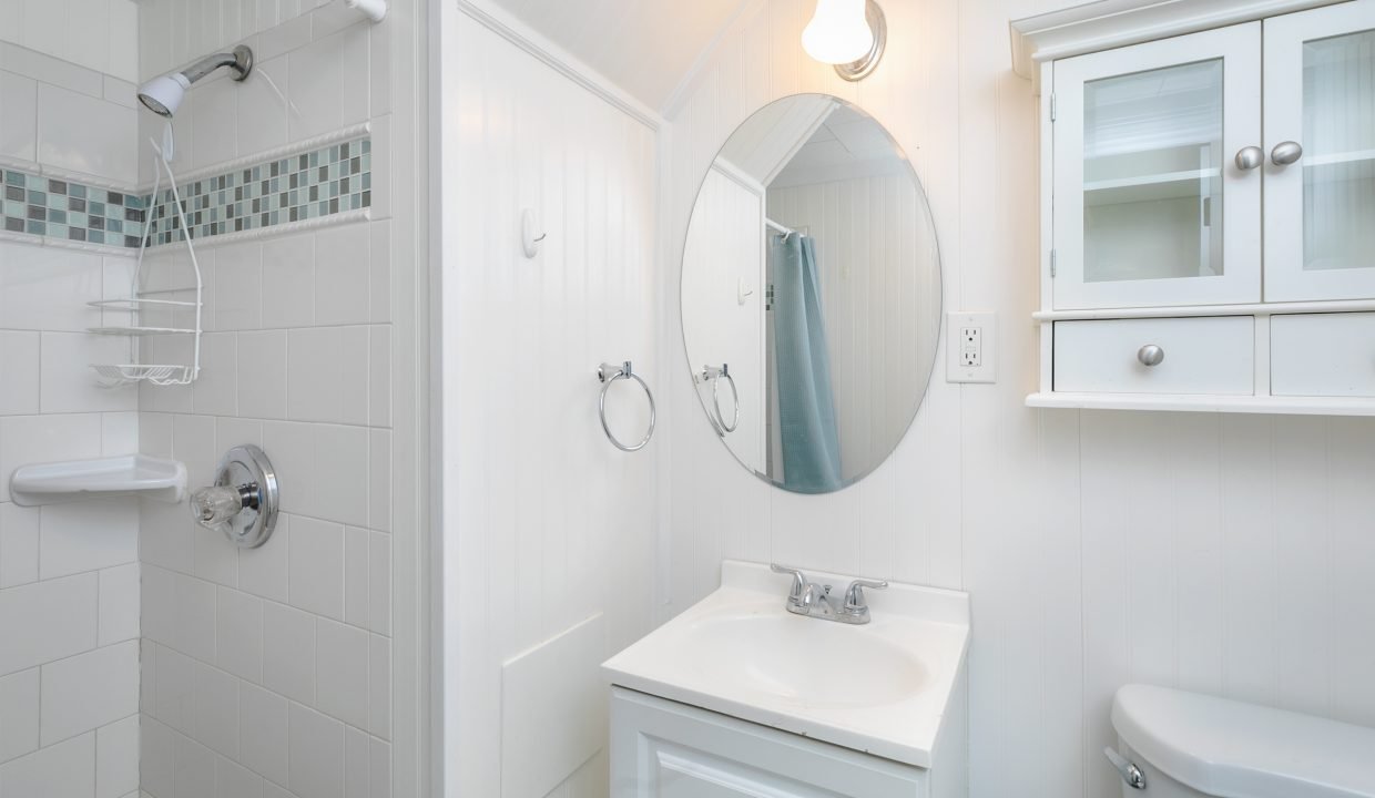 A small, bright bathroom with white walls, featuring a sink, mirror, and a shower area with a curtain rod.