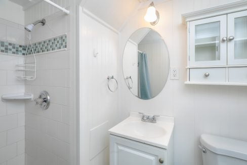 A small, bright bathroom with white walls, featuring a sink, mirror, and a shower area with a curtain rod.