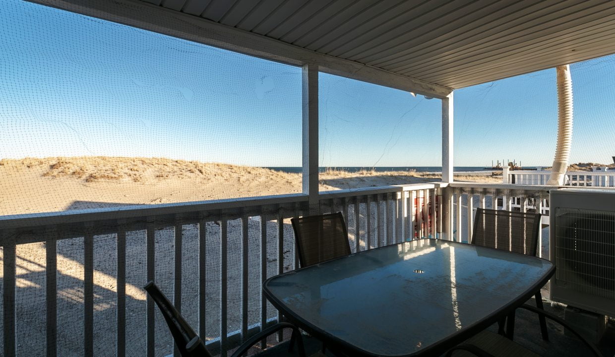 Screened porch with a table and chairs overlooking a sandy beach and clear blue sky.