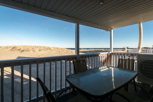 Screened porch with a table and chairs overlooking a sandy beach and clear blue sky.