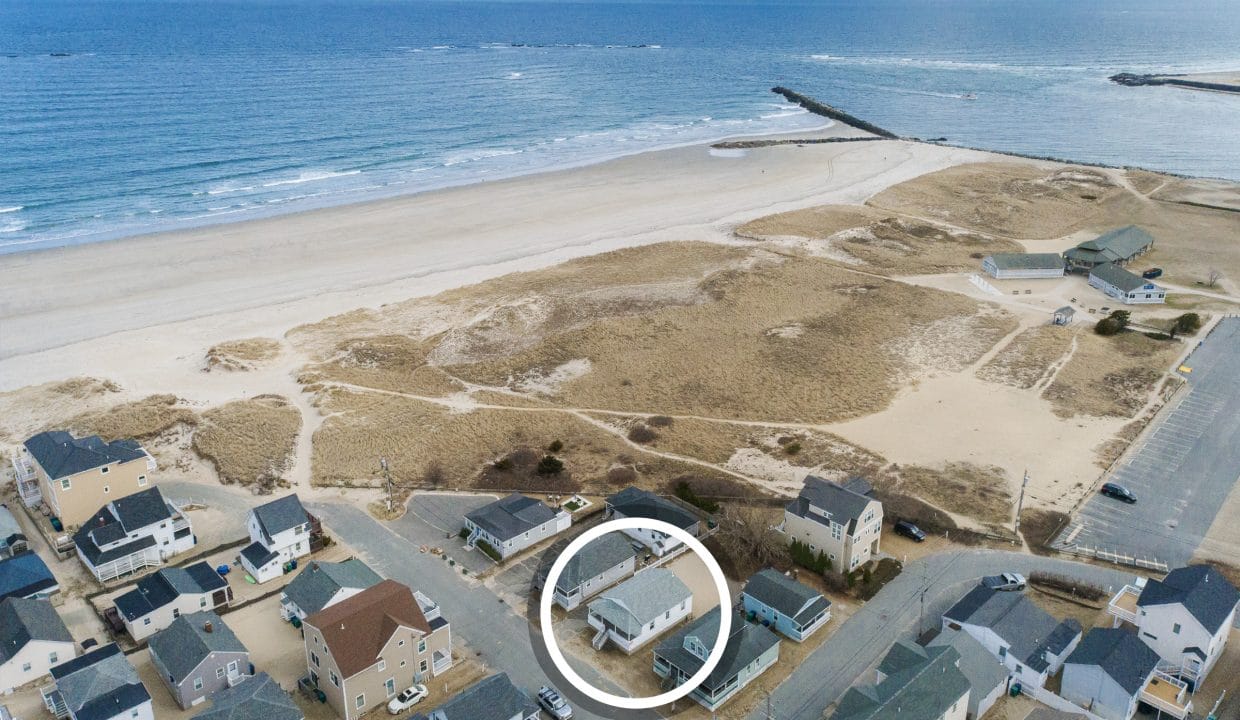Aerial view of beachfront houses and sandy dunes near the ocean, with a white circle highlighting one specific house in the foreground.
