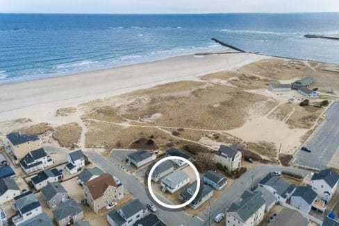 Aerial view of beachfront houses and sandy dunes near the ocean, with a white circle highlighting one specific house in the foreground.