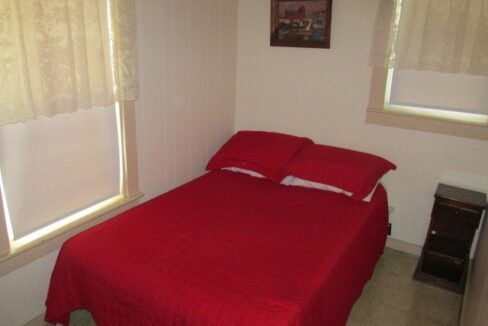 a bed with a red comforter and two windows.