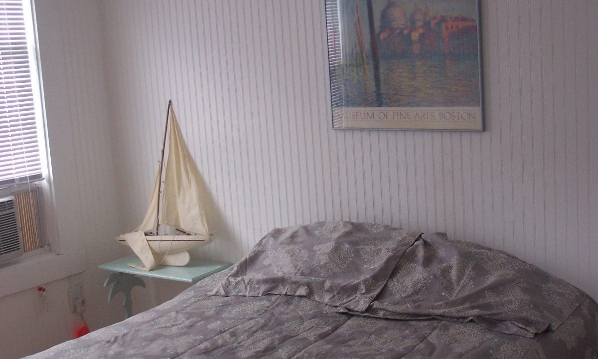 A neatly made bed with gray bedding next to a small table holding a model sailboat, under a framed 