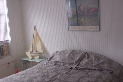 A neatly made bed with gray bedding next to a small table holding a model sailboat, under a framed 