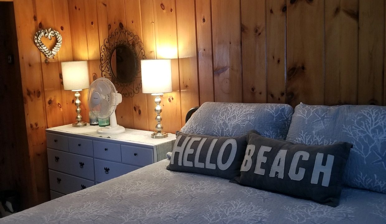A bed in a bedroom with wood paneling and a hello beach sign.