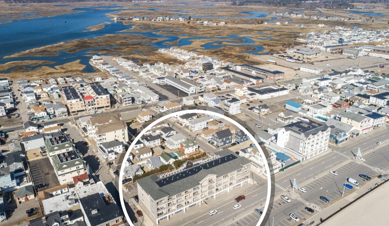 Aerial view of a coastal town with a large building near the beach and a parking lot, highlighted by a white circle in the foreground. Marshland and water are visible in the background.