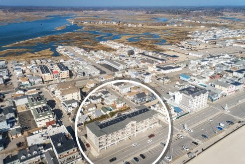 Aerial view of a coastal town with a large building near the beach and a parking lot, highlighted by a white circle in the foreground. Marshland and water are visible in the background.