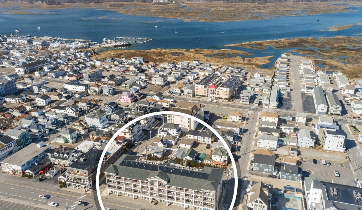 Aerial view of a coastal town with a large building in the foreground circled, showing nearby houses, roads, and marshy waterways in the background.