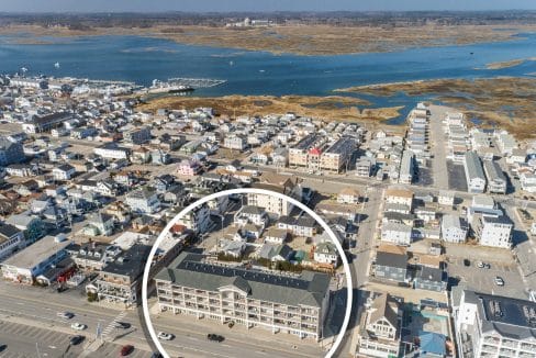 Aerial view of a coastal town with a large building in the foreground circled, showing nearby houses, roads, and marshy waterways in the background.