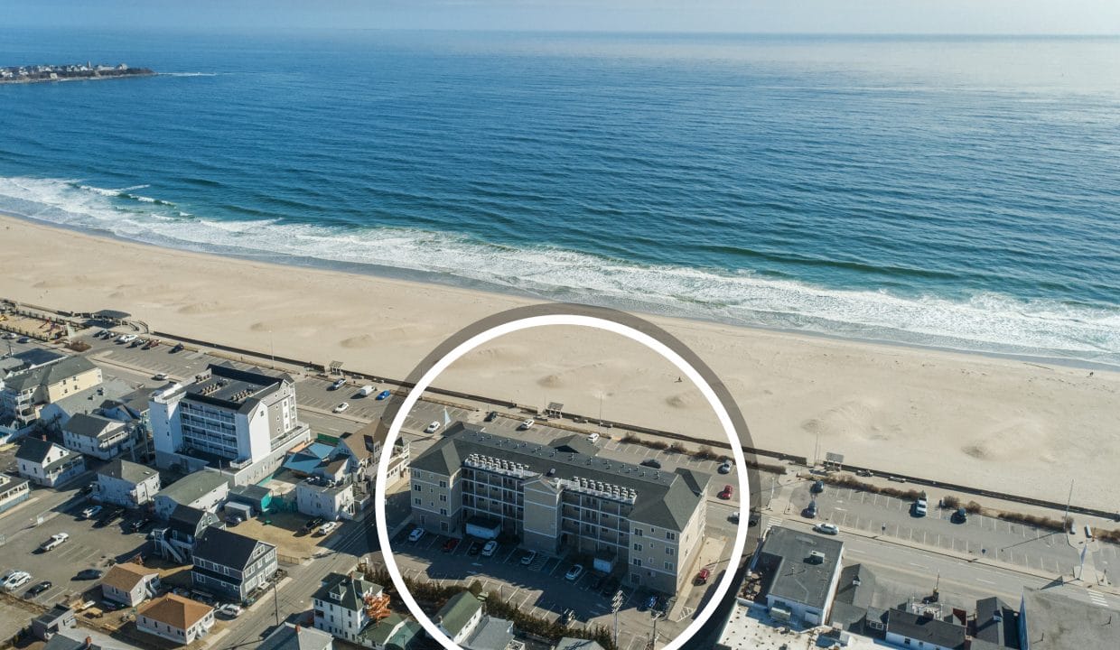Aerial view of a coastal town with a sandy beach and ocean; a specific building complex near the beach is highlighted with a white circle.