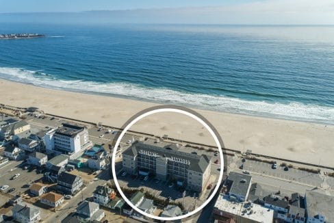 Aerial view of a coastal town with a sandy beach and ocean; a specific building complex near the beach is highlighted with a white circle.