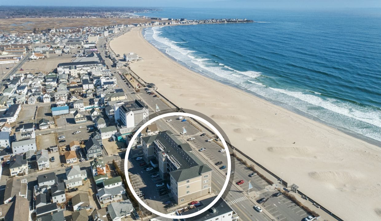 Aerial view of a coastal town with a sandy beach and ocean waves; a building near the shoreline is highlighted with a white circle.