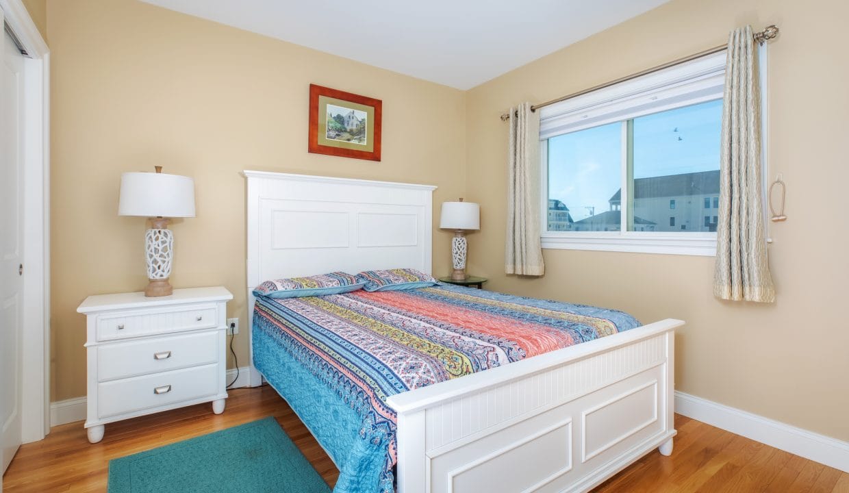 A bedroom with a white bed frame, colorful bedspread, two matching nightstands with lamps, a window with beige curtains, and a small framed picture on the wall.