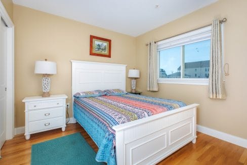 A bedroom with a white bed frame, colorful bedspread, two matching nightstands with lamps, a window with beige curtains, and a small framed picture on the wall.
