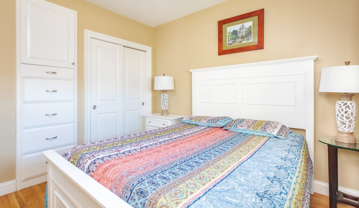 A neatly made bed with a colorful patterned bedspread in a beige bedroom, flanked by nightstands with lamps and a framed picture above the headboard.