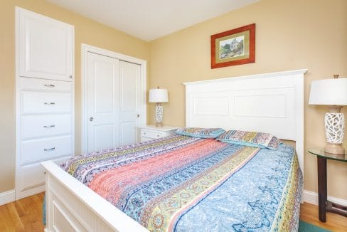 A neatly made bed with a colorful patterned bedspread in a beige bedroom, flanked by nightstands with lamps and a framed picture above the headboard.