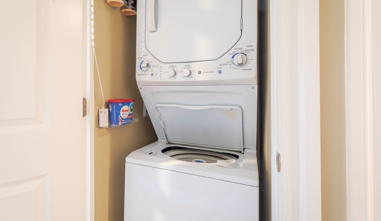 A stacked washer and dryer unit in a small laundry closet with the washer lid open and laundry supplies on a shelf nearby.