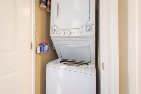 A stacked washer and dryer unit in a small laundry closet with the washer lid open and laundry supplies on a shelf nearby.