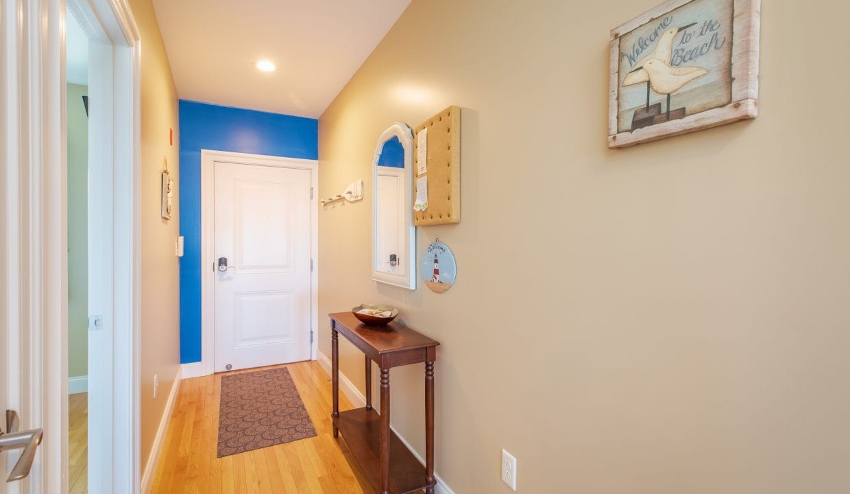 A hallway with tan walls, a wooden floor, a white door, a small console table, wall mirrors, and beach-themed decor including a framed sign that says 