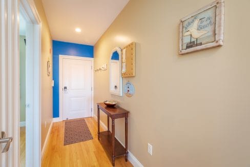 A hallway with tan walls, a wooden floor, a white door, a small console table, wall mirrors, and beach-themed decor including a framed sign that says 