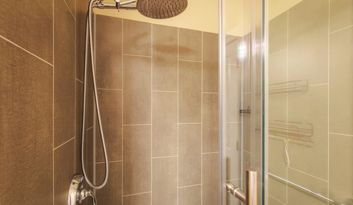 Modern shower with beige tile walls, a round rainfall showerhead, metal handle, glass door, and a metal shower caddy fixed to the wall.