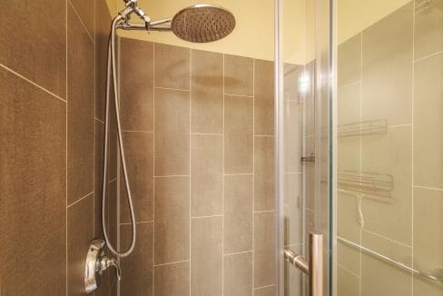 Modern shower with beige tile walls, a round rainfall showerhead, metal handle, glass door, and a metal shower caddy fixed to the wall.