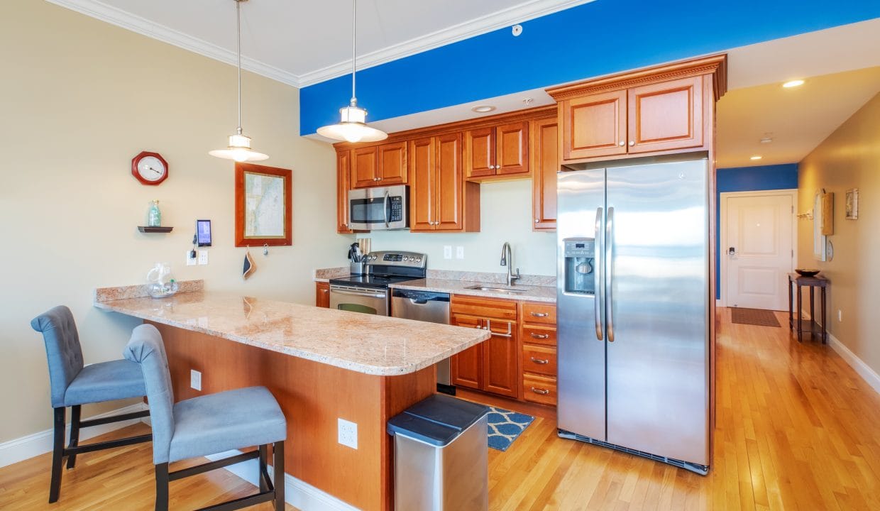 Modern kitchen with wooden cabinets, stainless steel appliances, granite countertops, two gray upholstered chairs at the breakfast bar, and light wood flooring.