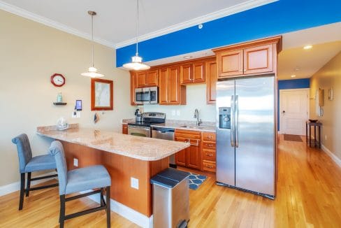 Modern kitchen with wooden cabinets, stainless steel appliances, granite countertops, two gray upholstered chairs at the breakfast bar, and light wood flooring.