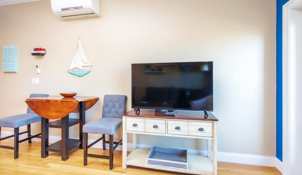 Flat-screen TV on a white console table in a living room with wooden floors, a small dining table with two chairs, and beige walls.