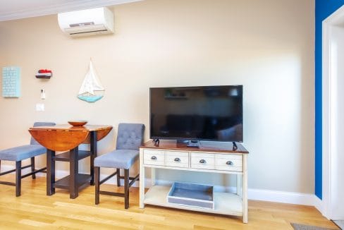 Flat-screen TV on a white console table in a living room with wooden floors, a small dining table with two chairs, and beige walls.