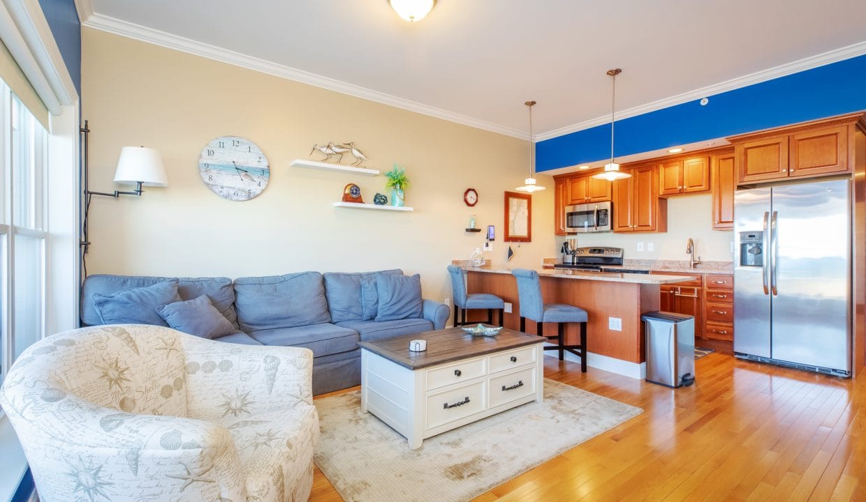 Bright, modern living room and kitchen with hardwood floors, gray sectional sofa, patterned armchair, and stainless steel appliances. Shelves and nautical decor on the wall.