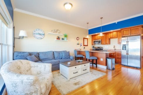 Bright, modern living room and kitchen with hardwood floors, gray sectional sofa, patterned armchair, and stainless steel appliances. Shelves and nautical decor on the wall.