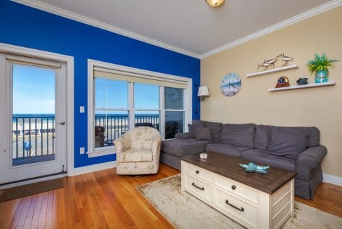 Living room with gray sectional sofa, armchair, and wooden coffee table; blue accent wall with windows and door showing ocean view, shelves with decor on beige wall.