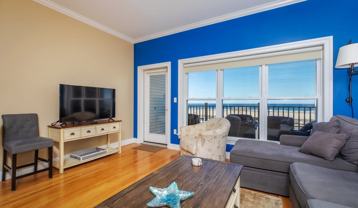 Living room with a blue accent wall, flat-screen TV on a stand, gray sectional sofa, armchair, coffee table, wooden floors, and large windows showing an ocean view.