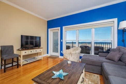 Living room with a blue accent wall, flat-screen TV on a stand, gray sectional sofa, armchair, coffee table, wooden floors, and large windows showing an ocean view.