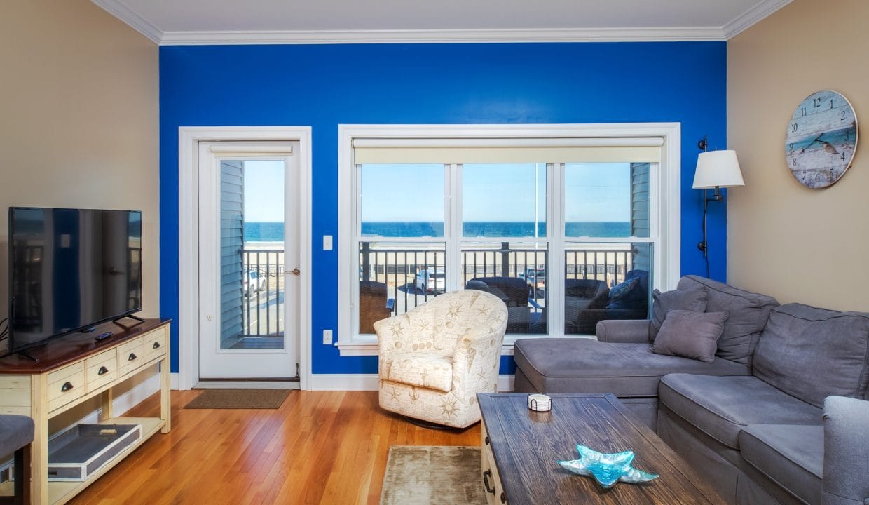 Living room with a blue accent wall, large window and glass door overlooking a beach, gray sectional sofa, armchair, TV, and wooden coffee table.
