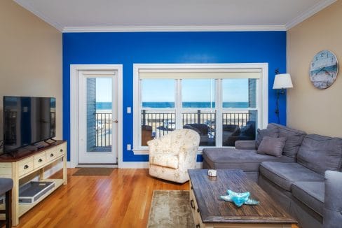Living room with a blue accent wall, large window and glass door overlooking a beach, gray sectional sofa, armchair, TV, and wooden coffee table.