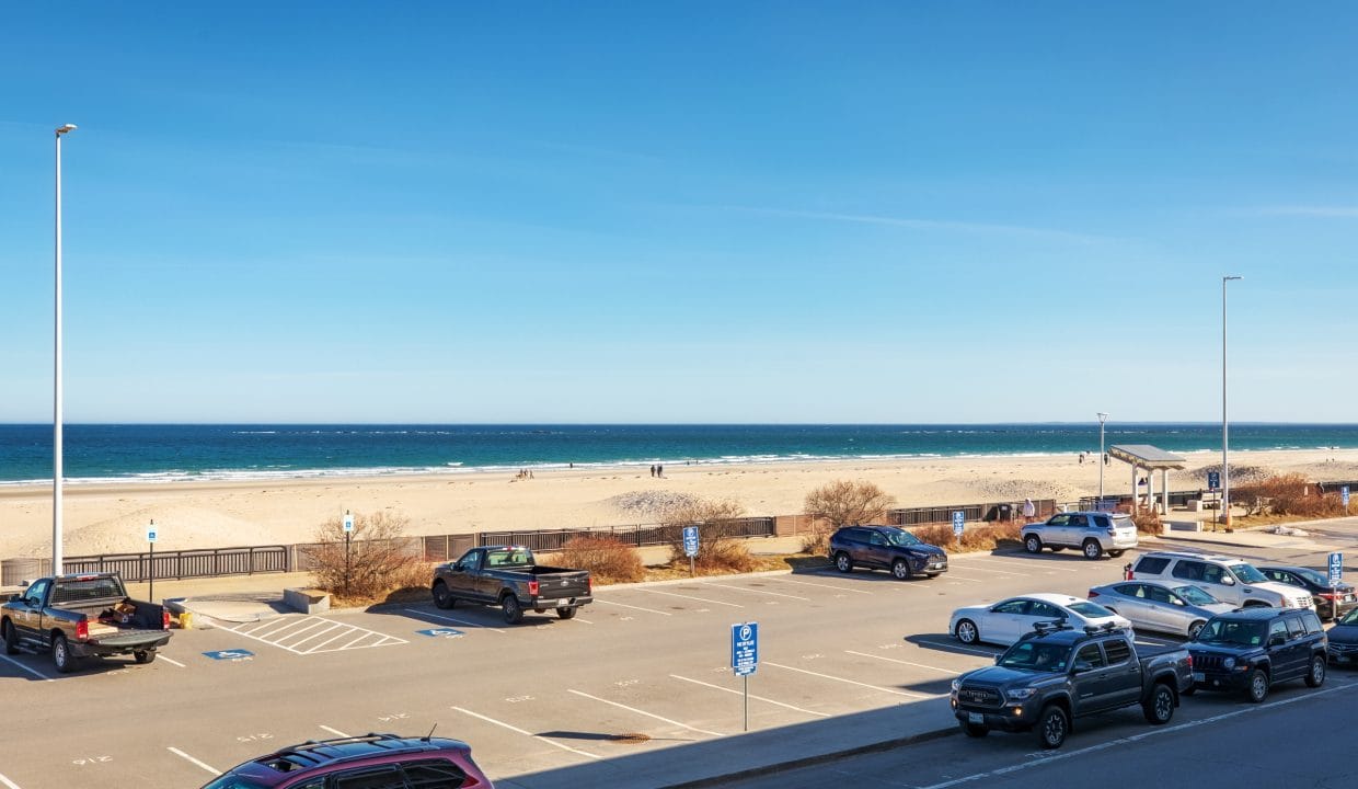A beach parking lot with several cars parked, a few people walking on the sandy beach, and the ocean visible under a clear blue sky.