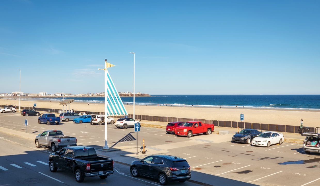 View of a beachside parking lot with several parked cars, a blue and white sail sculpture, sandy beach, and ocean under a clear blue sky.