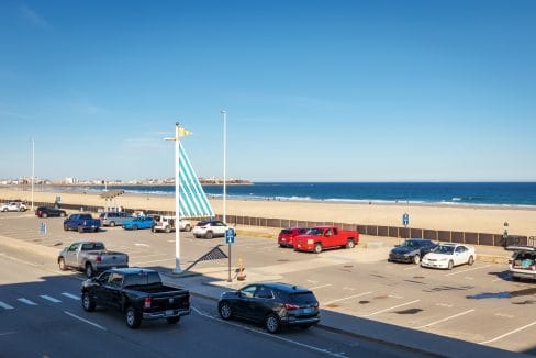 View of a beachside parking lot with several parked cars, a blue and white sail sculpture, sandy beach, and ocean under a clear blue sky.